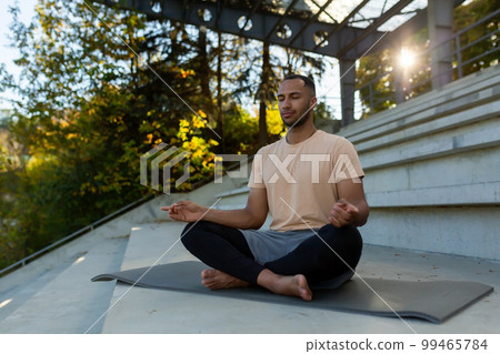 A young African American man is sitting on a mat in a stadium in the lotus position and doing yoga. He meditates, closed his eyes, relaxed, calm. 99465784
