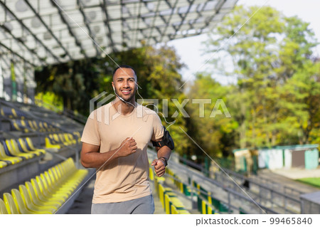 Young African American man, sportsman, track and field athlete running in headphones in the stadium between the stands in the summer. Smiling at the camera. 99465840