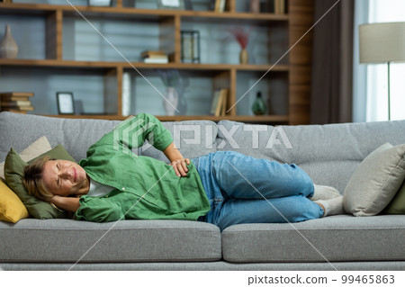 Mature woman alone at home sick, holding hands to side of stomach having severe spasm lying on sofa in living room. 99465863