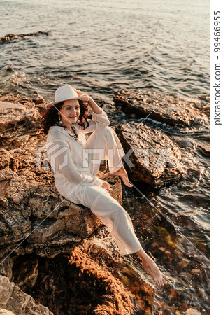 A woman in a white pantsuit and hat is standing on the beach enjoying the sea. Happy summer holidays 99466955