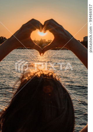 Silhouette against the sunset - hands folded in the shape of a heart. Sun with rays between the palms. Concept for Valentine's Day, declaration of love. Minimalism, space for text, warm colors. 99466957