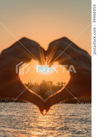 Silhouette against the sunset - hands folded in the shape of a heart. Sun with rays between the palms. Concept for Valentine's Day, declaration of love. Minimalism, space for text, warm colors. Silhouette against the sunset - hands folded in the shape of a heart. Sun with rays between the palms. Concept for Valentine's Day, declaration of love. Minimalism, space for text, warm colors. 99466959