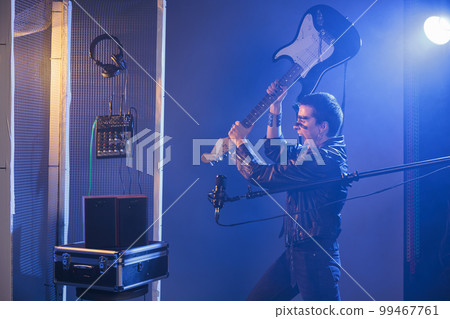 Crazy funky rocker throwing guitar in studio, wearing leather jacket and having cool make up in studio. Guitarist acting playful and silly, smashing instrument and singing punk rock music. Crazy funky rocker throwing guitar in studio, wearing leather jacket and having cool make up in studio. Guitarist acting playful and silly, smashing instrument and singing punk rock music. 99467761