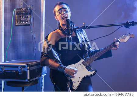Talented guitarist singing alternative rock at microphone, playing electric guitar in studio. Doing live heavy metal performance and feeling ecstatic, screaming loud and enjoying solo concert. Talented guitarist singing alternative rock at microphone, playing electric guitar in studio. Doing live heavy metal performance and feeling ecstatic, screaming loud and enjoying solo concert. 99467783