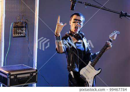 Musical artist doing rock sign and playing guitar, feeling ecstatic and doing solo concert in studio over background. Female singer showing rock and roll symbol at live show performance. 99467784