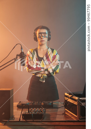 Woman artist posing with arms crossed at turntables, using headphones and mixer to produce techno music or sounds. Doing performance with mixer and control buttons, audio stereo equipment. 99467795