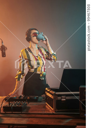 Young woman DJ drinking coffee from mug before mixing techno music, preparing to do performance on turntables. Holding caffeine drink in cup and doing remix with volume mixer. 99467808
