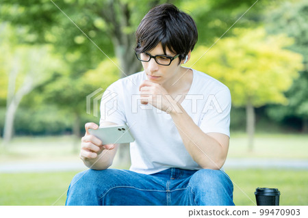 A man with glasses watching a smartphone video in a fresh green park 99470903