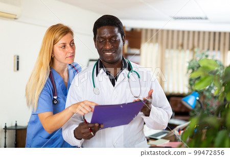 Two multinational doctors in the clinic discuss work issues while standing in an office 99472856