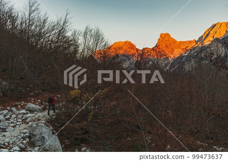 Man in red hood hiking the trail toward the alps in sunset. 99473637