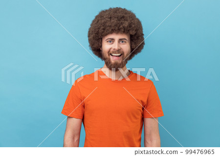Portrait of man wearing orange T-shirt looking at camera with toothy smile and happy facial expression, being in good mood, rejoicing great results. Indoor studio shot isolated on blue background. Portrait of man wearing orange T-shirt looking at camera with toothy smile and happy facial expression, being in good mood, rejoicing great results. Indoor studio shot isolated on blue background. 99476965