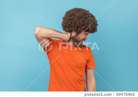 Portrait of man with Afro hairstyle in T-shirt touching neck, feeling acute pain moving and turning head, suffering spine problems, osteochondrosis. Indoor studio shot isolated on blue background. 99476968
