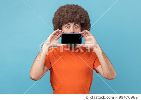 Portrait of funny positive man with Afro hairstyle in orange T-shirt covering mouth with smart phone with empty display, looking at camera with big eyes. Indoor studio shot isolated on blue background 99476969