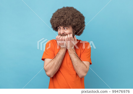 Anxiety disorder. Portrait of man with Afro hairstyle wearing orange T-shirt biting nails, nervous about troubles, panicking and looking scared. Indoor studio shot isolated on blue background. Anxiety disorder. Portrait of man with Afro hairstyle wearing orange T-shirt biting nails, nervous about troubles, panicking and looking scared. Indoor studio shot isolated on blue background. 99476976