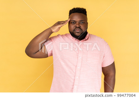Yes sir. Portrait of serious man wearing pink shirt holding hand near head as soldier and saluting, looking attentive and confident. Indoor studio shot isolated on yellow background. 99477004