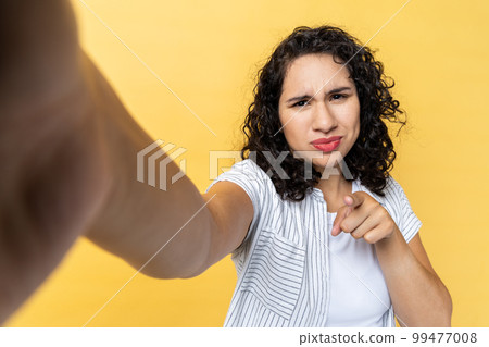 Beautiful young adult woman blogger with dark wavy hair making point of view photo or broadcasting livestream, pointing finger to camera, POV. Indoor studio shot isolated on yellow background. 99477008