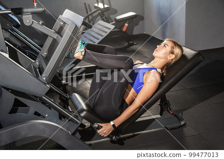 Side view portrait of strong powerful woman doing fitness training on a leg extension, push machine with weights in a gym, wearing wearing sports top and tights. Indoor shot. 99477013