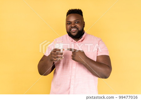 Portrait of smiling satisfied bearded man wearing pink shirt holding glass of water with ice, pointing at cold beverage, looking at camera. Indoor studio shot isolated on yellow background. 99477016