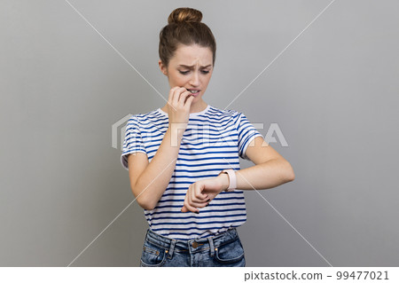 I need more time. Portrait of nervous woman wearing striped T-shirt standing and looking at her smart watch and want more time, biting fingernails. Indoor studio shot isolated on gray background. 99477021