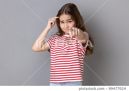 You're idiot with insane crazy plan. Little girl wearing striped T-shirt showing stupid gesture and pointing to camera, accusing of dumb idea. Indoor studio shot isolated on gray background. You're idiot with insane crazy plan. Little girl wearing striped T-shirt showing stupid gesture and pointing to camera, accusing of dumb idea. Indoor studio shot isolated on gray background. 99477024