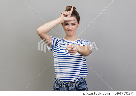 Portrait of woman wearing striped T-shirt expressing disrespect, showing L finger sign to camera, loser gesture, accusing for failure. Indoor studio shot isolated on gray background. 99477037