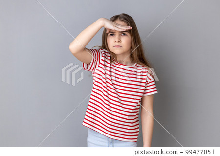 Portrait of little girl wearing striped T-shirt holding palm over her eyes, looking into distance with attentive view, watching horizon, far future. Indoor studio shot isolated on gray background. Portrait of little girl wearing striped T-shirt holding palm over her eyes, looking into distance with attentive view, watching horizon, far future. Indoor studio shot isolated on gray background. 99477051