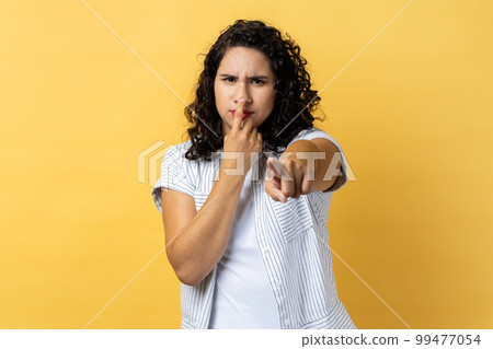 I don't believe, you are liar. Dissatisfied woman with dark wavy hair in business suit touching nose showing lie gesture, suspecting cheating. Indoor studio shot isolated on yellow background. 99477054