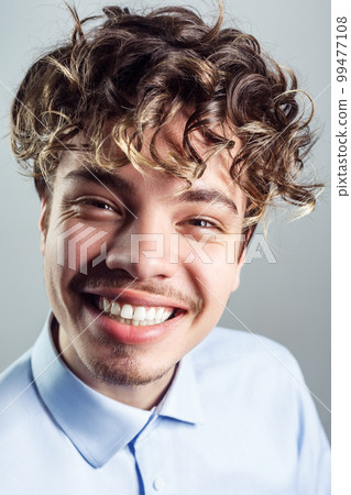 Portrait of happy smiling handsome man with mustache with curly hairstyle, wearing blue shirt, looking at camera, expressing happiness. Indoor studio shot isolated on gray background. Portrait of happy smiling handsome man with mustache with curly hairstyle, wearing blue shirt, looking at camera, expressing happiness. Indoor studio shot isolated on gray background. 99477108