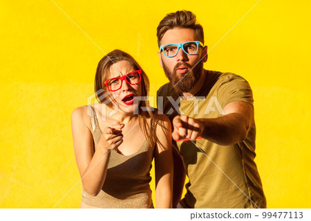 Portrait of astonished amazed man and woman in glasses standing with shocked face and pointing to camera, having surprised expression. Indoor studio shot isolated on yellow background. Portrait of astonished amazed man and woman in glasses standing with shocked face and pointing to camera, having surprised expression. Indoor studio shot isolated on yellow background. 99477113