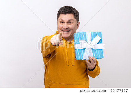 Portrait of satisfied middle aged handsome man standing, holding present box, pointing finger to camera, wearing urban style hoodie. Indoor studio shot isolated on white background. 99477157