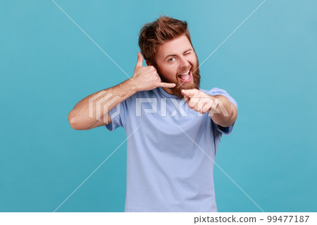 You call me. Portrait of playful bearded man flirting and pointing at camera with attractive smile, making call me gesture with fingers. Indoor studio shot isolated on blue background. 99477187