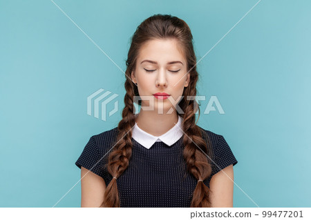 Portrait of attractive beautiful calm woman with braids standing with closed eyes, having problems with eyesight, wearing black dress. woman Indoor studio shot isolated on blue background. 99477201