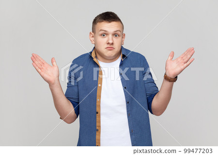 I don't know, who cares. Portrait of confused clueless teenager boy wearing blue shirt shrugging shoulders, making no idea gesture, whatever. Indoor studio shot isolated on gray background. I don't know, who cares. Portrait of confused clueless teenager boy wearing blue shirt shrugging shoulders, making no idea gesture, whatever. Indoor studio shot isolated on gray background. 99477203