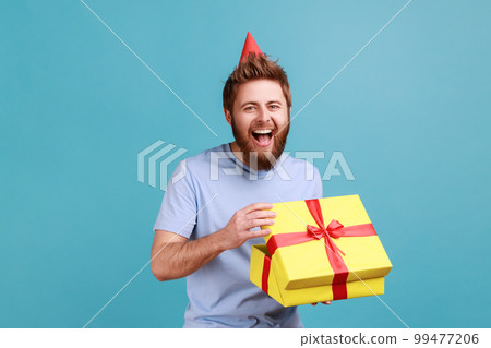 Man with party cone hat holding opened gift box and looking at camera with happy smile, unpacking present, enjoying awesome birthday surprise. Indoor studio shot isolated on blue background. 99477206