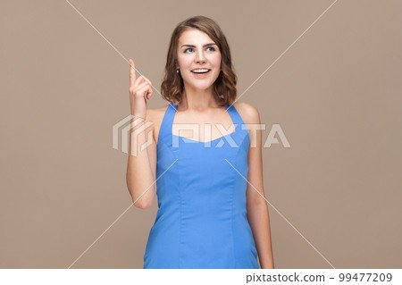 Portrait of overjoyed excited woman with wavy hair raises fore finger as receives good idea, exclaims with amazement, wearing blue dress. Indoor studio shot isolated on light brown background. 99477209