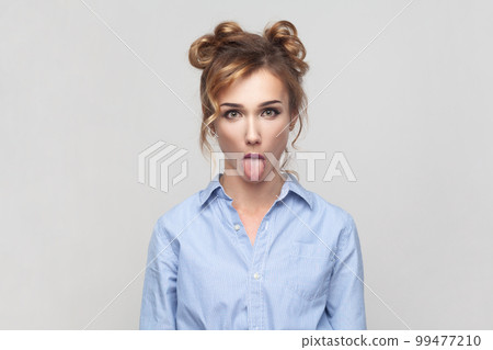 Portrait of naughty comic blonde woman sticking out tongue, misbehaves, demonstrating her disobedience and protest, has funny look, wearing blue shirt. Indoor studio shot isolated on gray background. 99477210