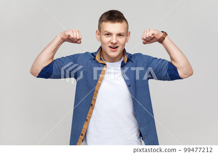 Portrait of strong teenager boy wearing blue shirt demonstrating power in his hands, showing biceps, feeling energy to win success. Indoor studio shot isolated on gray background. Portrait of strong teenager boy wearing blue shirt demonstrating power in his hands, showing biceps, feeling energy to win success. Indoor studio shot isolated on gray background. 99477212
