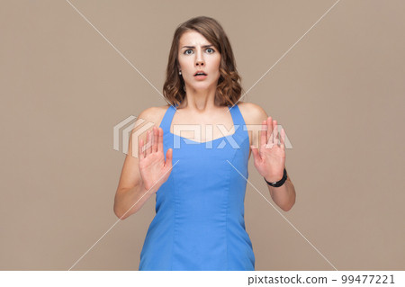 Portrait of shocked frighten young adult woman with wavy hair raised palms, showing stop gesture, sees something scary, wearing blue dress. Indoor studio shot isolated on light brown background. Portrait of shocked frighten young adult woman with wavy hair raised palms, showing stop gesture, sees something scary, wearing blue dress. Indoor studio shot isolated on light brown background. 99477221
