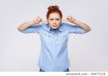 Don't want to listen. Portrait of annoyed redhead woman wearing blue shirt irritated by loud noise covering ears and grimacing in pain. Indoor studio shot isolated on gray background. Don't want to listen. Portrait of annoyed redhead woman wearing blue shirt irritated by loud noise covering ears and grimacing in pain. Indoor studio shot isolated on gray background. 99477229