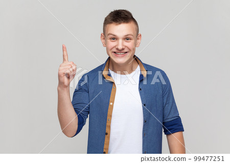 Portrait of excited amazed teenager boy wearing blue shirt pointing finger up with genius idea, surprised by suddenly invented smart solution. Indoor studio shot isolated on gray background. Portrait of excited amazed teenager boy wearing blue shirt pointing finger up with genius idea, surprised by suddenly invented smart solution. Indoor studio shot isolated on gray background. 99477251