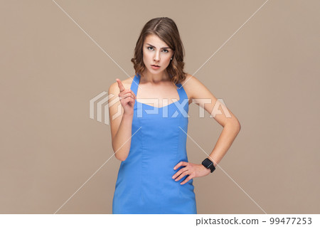 Portrait of serious angry young adult woman with wavy hair standing with raised index finger, warning you, wearing blue dress. Indoor studio shot isolated on light brown background. Portrait of serious angry young adult woman with wavy hair standing with raised index finger, warning you, wearing blue dress. Indoor studio shot isolated on light brown background. 99477253