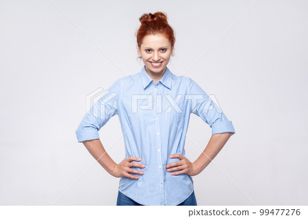 Portrait of positive happy pretty redhead woman wearing blue shirt standing with with hands on hips, looking at camera with toothy smile. Indoor studio shot isolated on gray background. 99477276