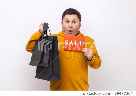Portrait of shocked man holding paper bags and sale sign, shopping and discounts, looking at camera with open mouth, wearing urban style hoodie. Indoor studio shot isolated on white background. Portrait of shocked man holding paper bags and sale sign, shopping and discounts, looking at camera with open mouth, wearing urban style hoodie. Indoor studio shot isolated on white background. 99477304