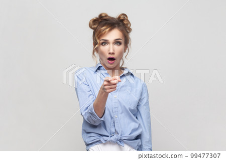 Portrait of surprised startled emotional blonde woman pointing fore finger straight at camera, opens mouth from shock, demonstrates something in front. Indoor studio shot isolated on gray background. Portrait of surprised startled emotional blonde woman pointing fore finger straight at camera, opens mouth from shock, demonstrates something in front. Indoor studio shot isolated on gray background. 99477307