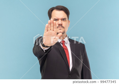 Portrait of strict bossy man with mustache showing block gesture, trying to stop conflict, looking at camera, wearing black suit with red tie. Indoor studio shot isolated on light blue background. 99477345