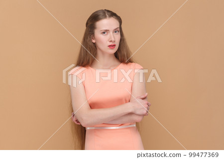 Portrait of pensive beautiful woman with long hair standing with with folded hands, looking at camera with thoughtful, wearing elegant dress. Indoor studio shot isolated on brown background. Portrait of pensive beautiful woman with long hair standing with with folded hands, looking at camera with thoughtful, wearing elegant dress. Indoor studio shot isolated on brown background. 99477369