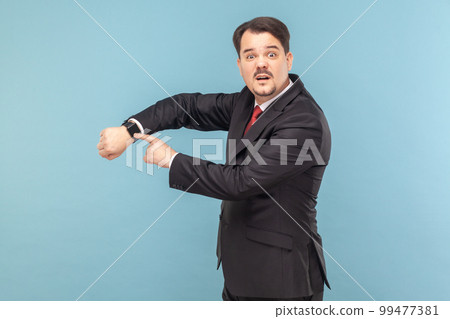 Confused man with mustache standing and looking at camera with puzzled expression, pointing at his wristwatch, wearing black suit with red tie. Indoor studio shot isolated on light blue background. 99477381