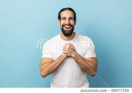 Portrait of satisfied delighted handsome man with beard wearing white T-shirt standing with hands together, looking at camera with amazed positive face. Indoor studio shot isolated on blue background. Portrait of satisfied delighted handsome man with beard wearing white T-shirt standing with hands together, looking at camera with amazed positive face. Indoor studio shot isolated on blue background. 99477399