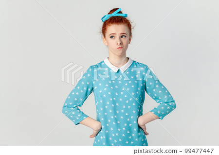 Portrait of confused puzzled ginger woman standing with hands on hips, looking away, thinking, keeps hands on hips, wearing blue dress. Indoor studio shot isolated on gray background. 99477447