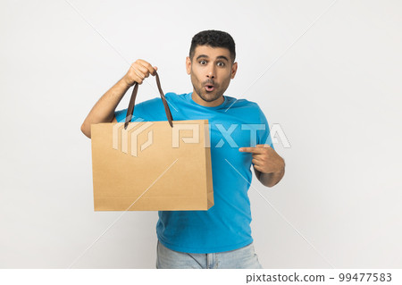 Portrait of shocked surprised astonished unshaven man wearing blue T- shirt standing pointing at shopping bag, great purchase in mall. Indoor studio shot isolated on gray background. 99477583